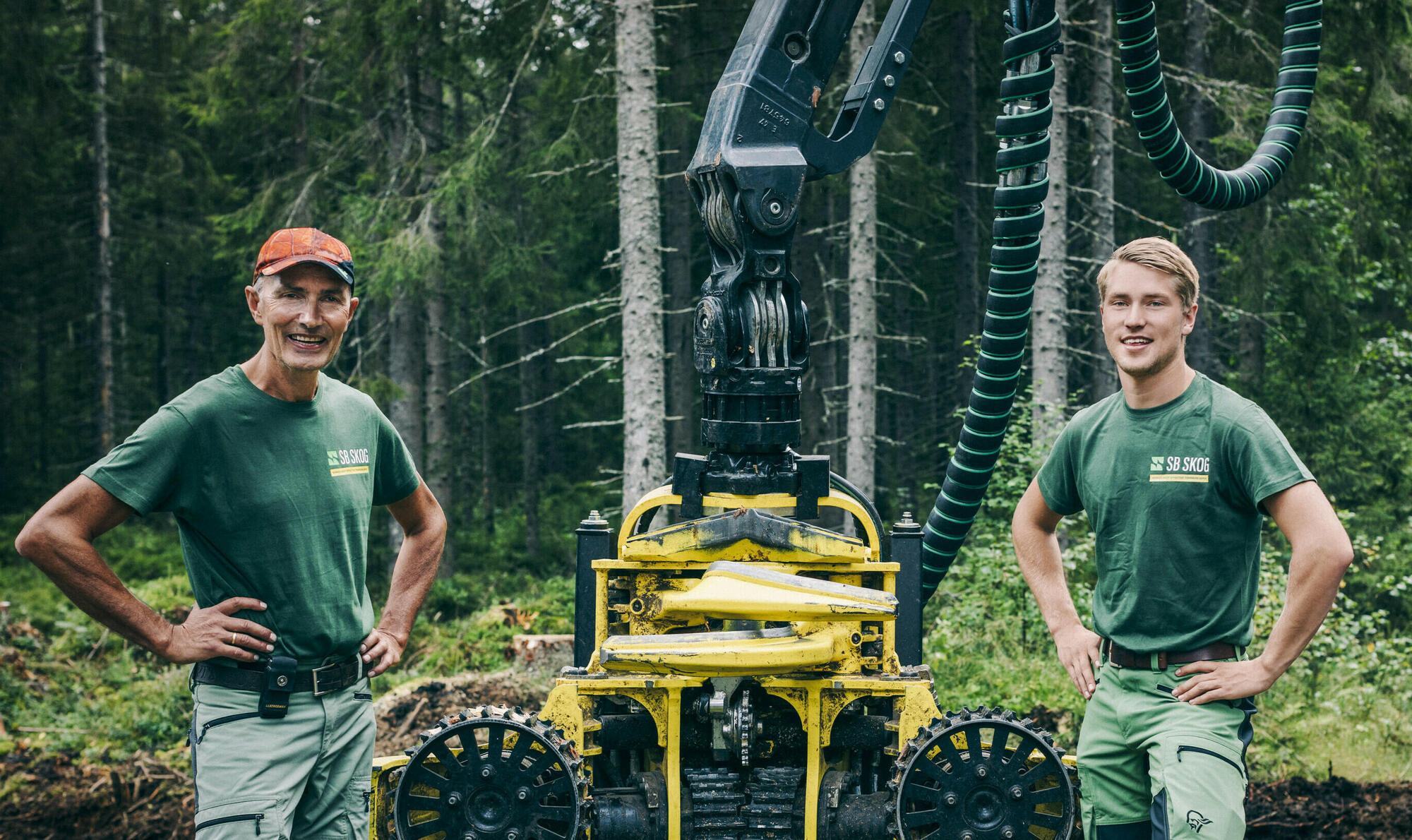 To SB Skog ansatte iført grønne t-skjorter står foran en gul hogstmaskin i frodig skog.
