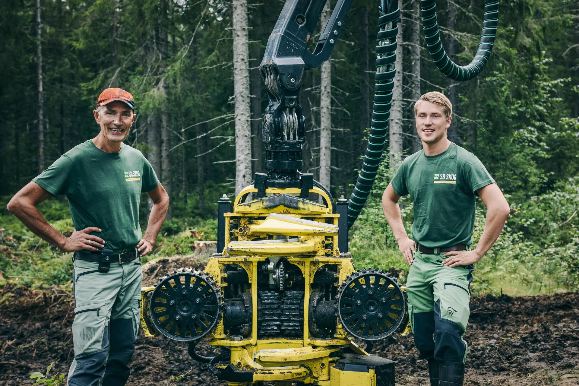 To SB Skog ansatte if&oslash;rt gr&oslash;nne t-skjorter st&aring;r foran en gul hogstmaskin i frodig skog.